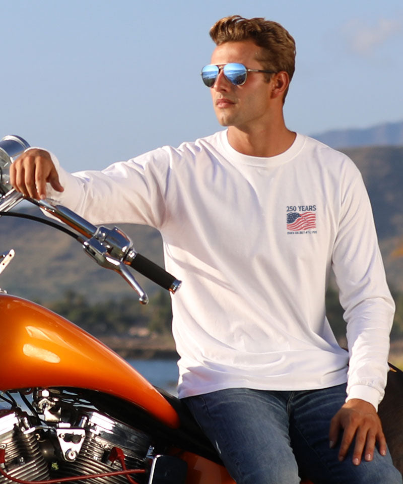 Man wearing a white long-sleeve shirt with a logo, sitting on an orange motorcycle with mountains in the background. View 3
