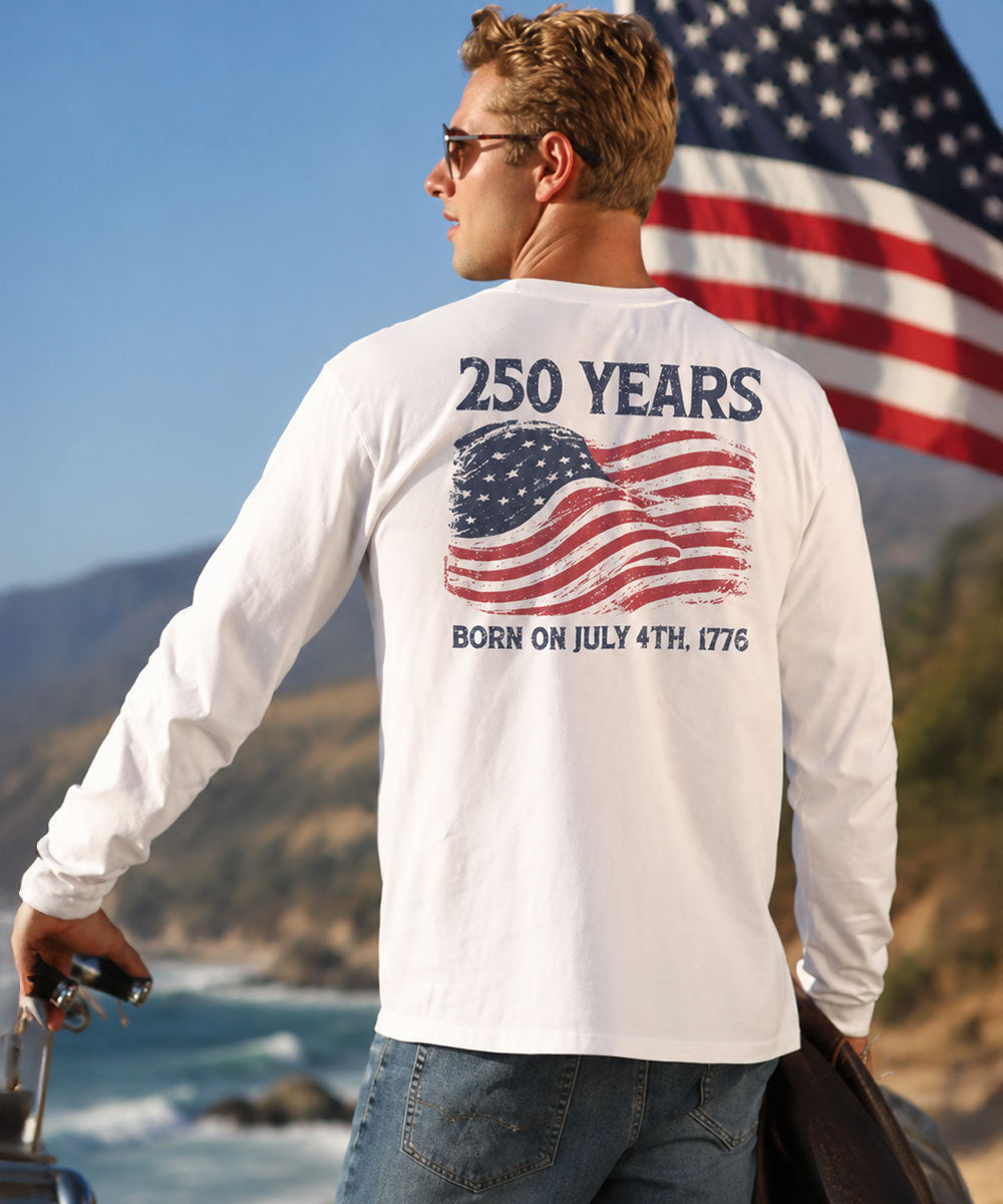 Man wearing a white long-sleeve shirt with '250 Years' and American flag design, standing in front of an American flag and mountains.