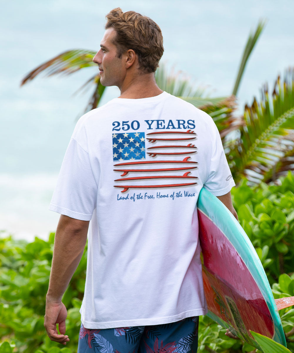 Man holding a surfboard with a white t-shirt featuring a graphic design in a tropical setting View 2