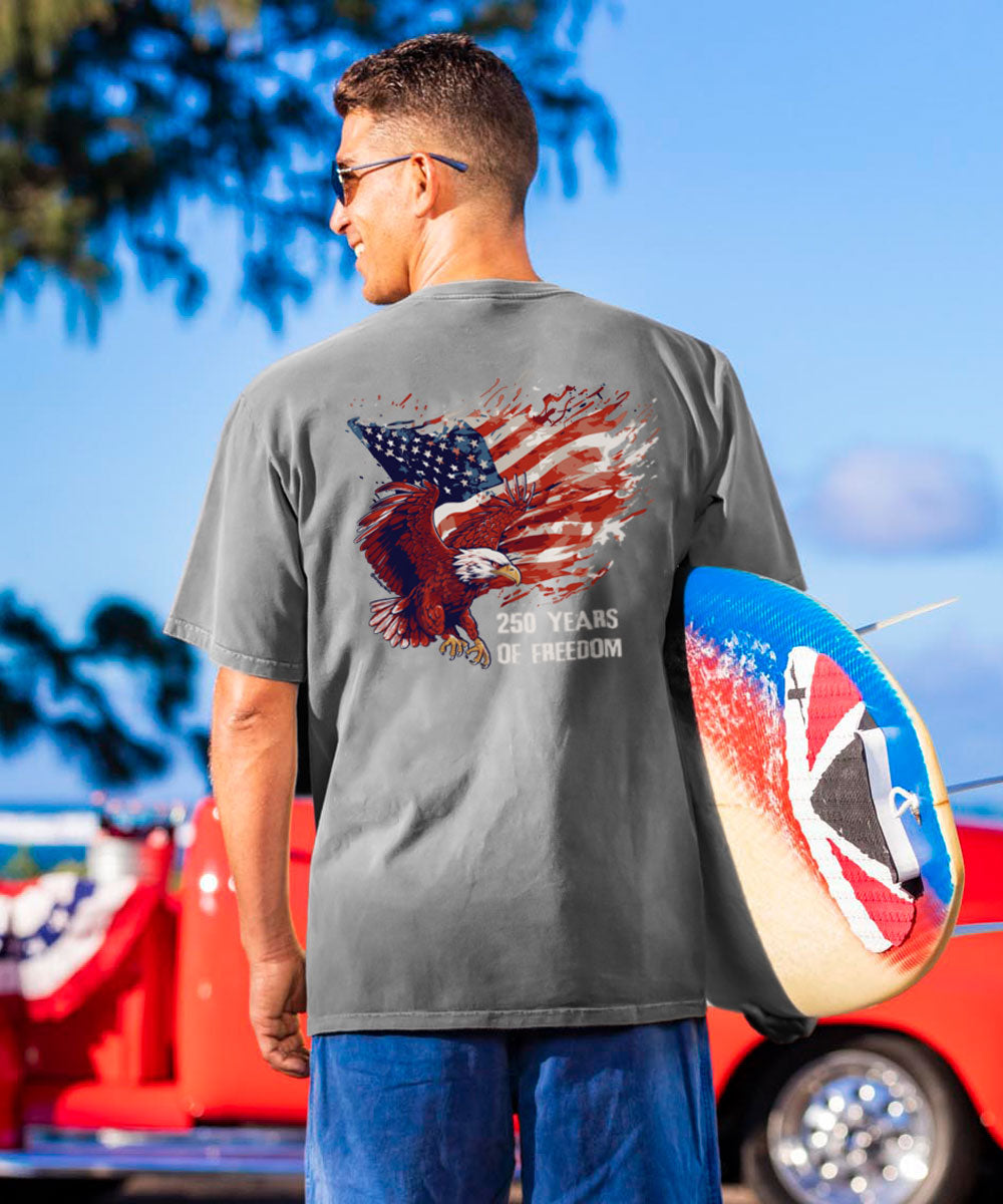 Man wearing a gray t-shirt with an eagle and American flag design, holding a surfboard with a Union Jack design, against a blue sky.