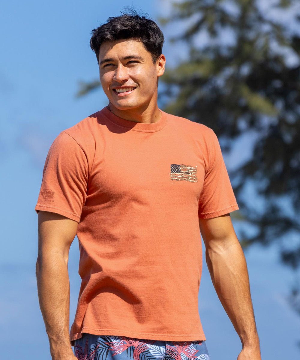 Man wearing an orange t-shirt with a logo, standing outdoors against a blue sky. View 3