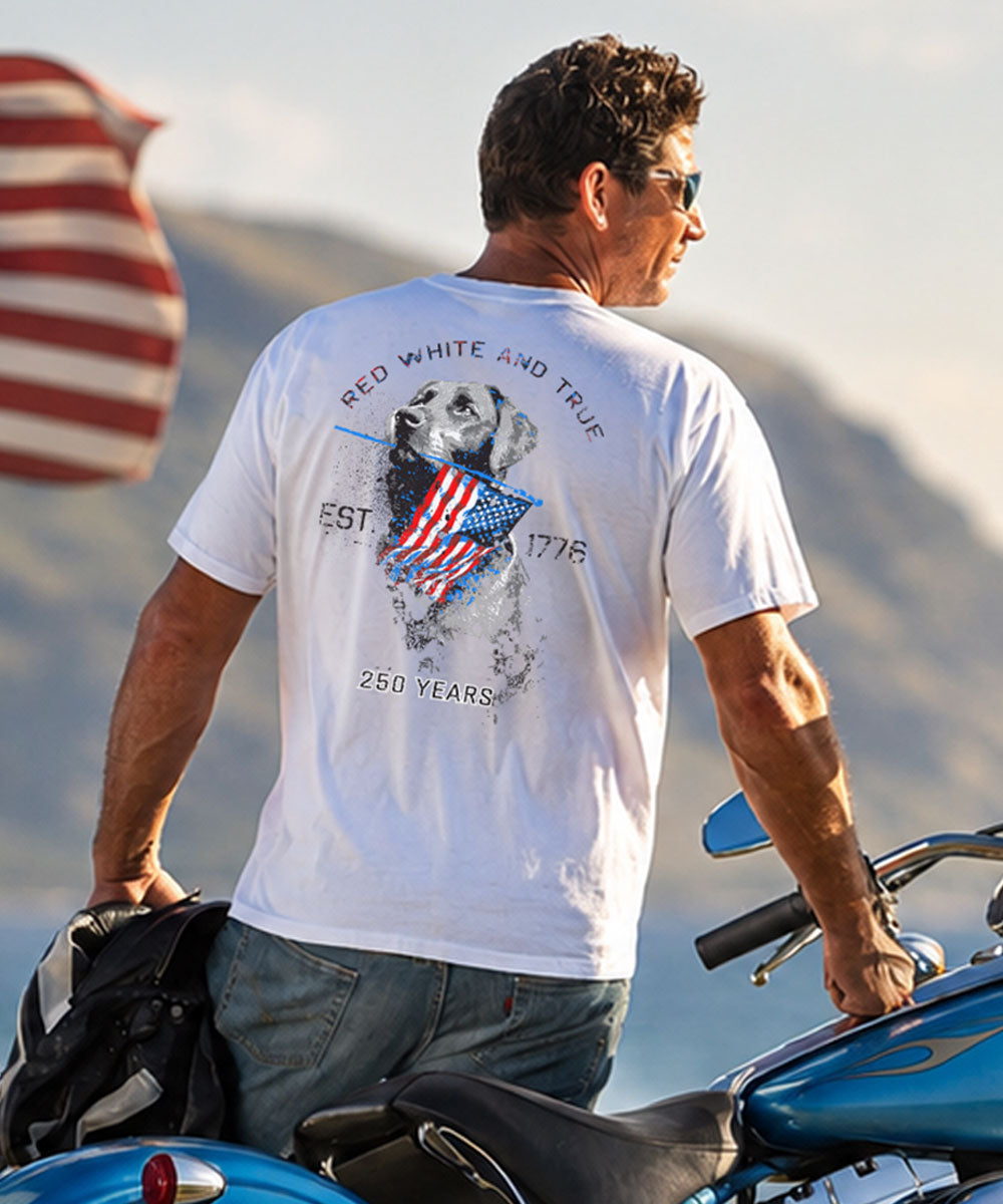 Man wearing a white t-shirt with a graphic design, standing next to a motorcycle with mountains in the background. View 2