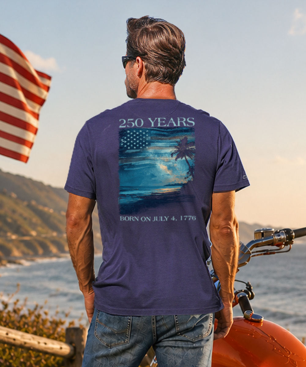 Man wearing a blue t-shirt with a sunset design and text, standing by a body of water with an American flag. View 2