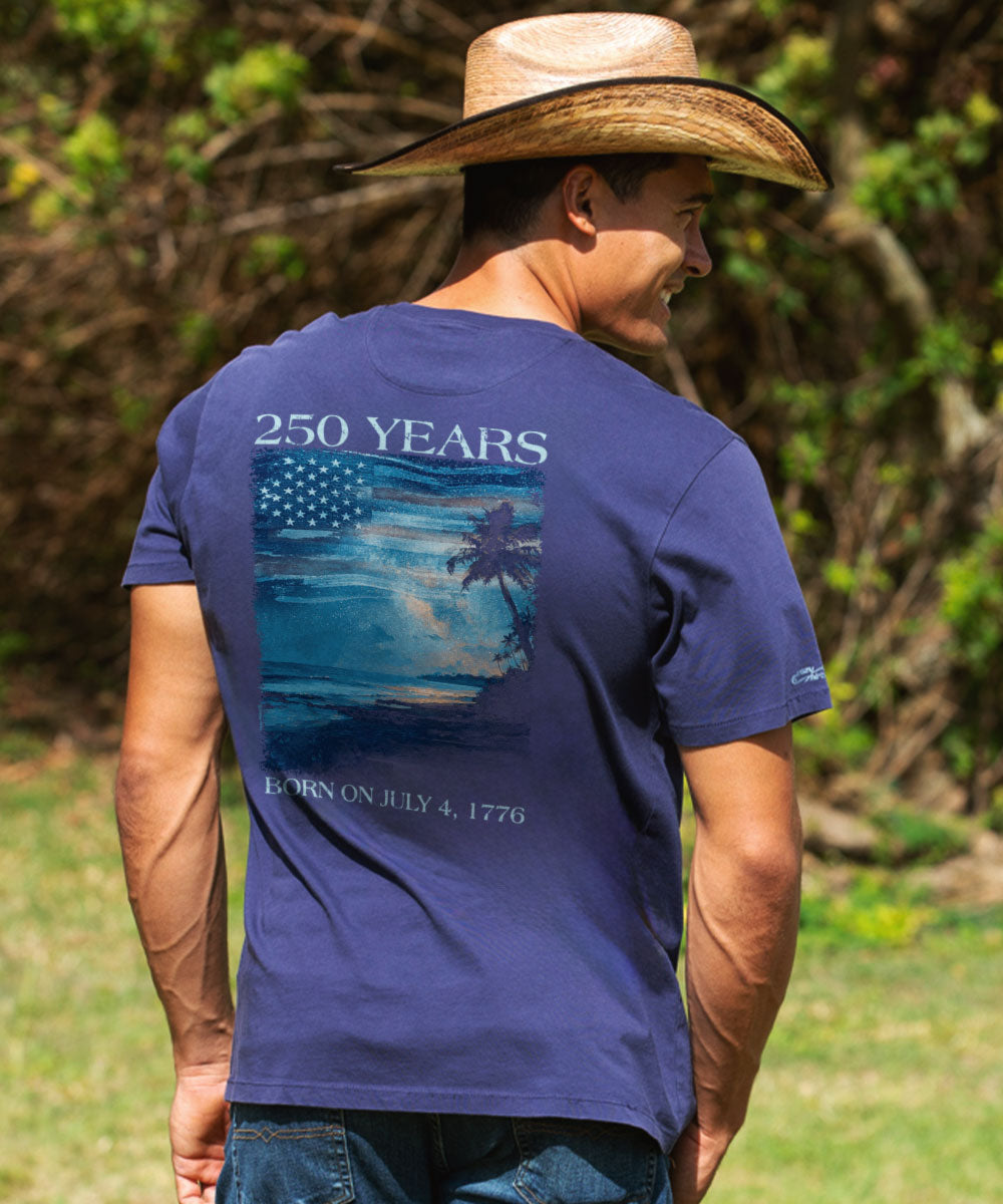 Man wearing a blue t-shirt with a graphic design and text, standing outdoors. View 2