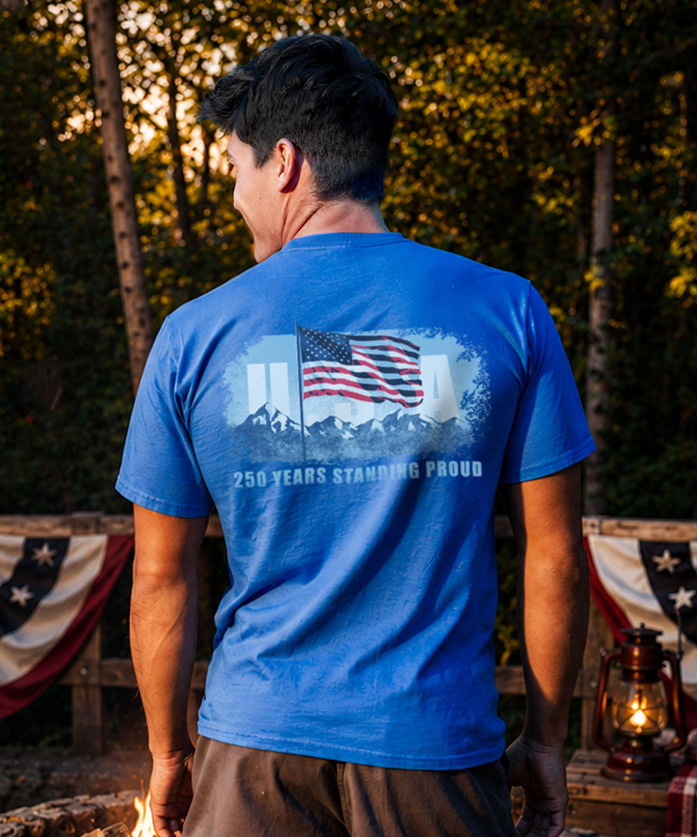 Person wearing a blue t-shirt with an American flag design and text, standing outdoors with trees and flags in the background.
