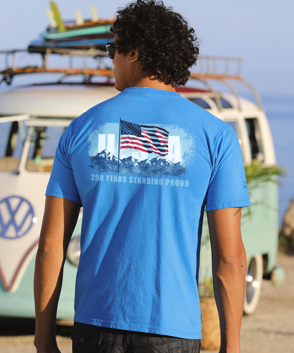 Person wearing a blue t-shirt with an American flag graphic and text, standing in front of a vintage van.