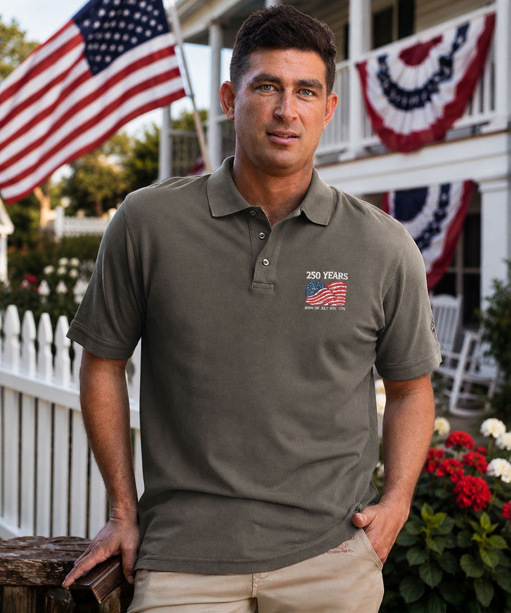 Man wearing a gray polo shirt with a 250th anniversary design, standing in front of a house with American flags. View 2