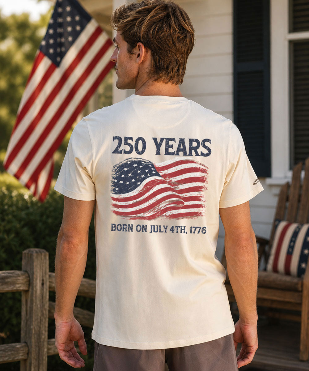 Person wearing a t-shirt with '250 Years' and American flag design, standing in front of an American flag.