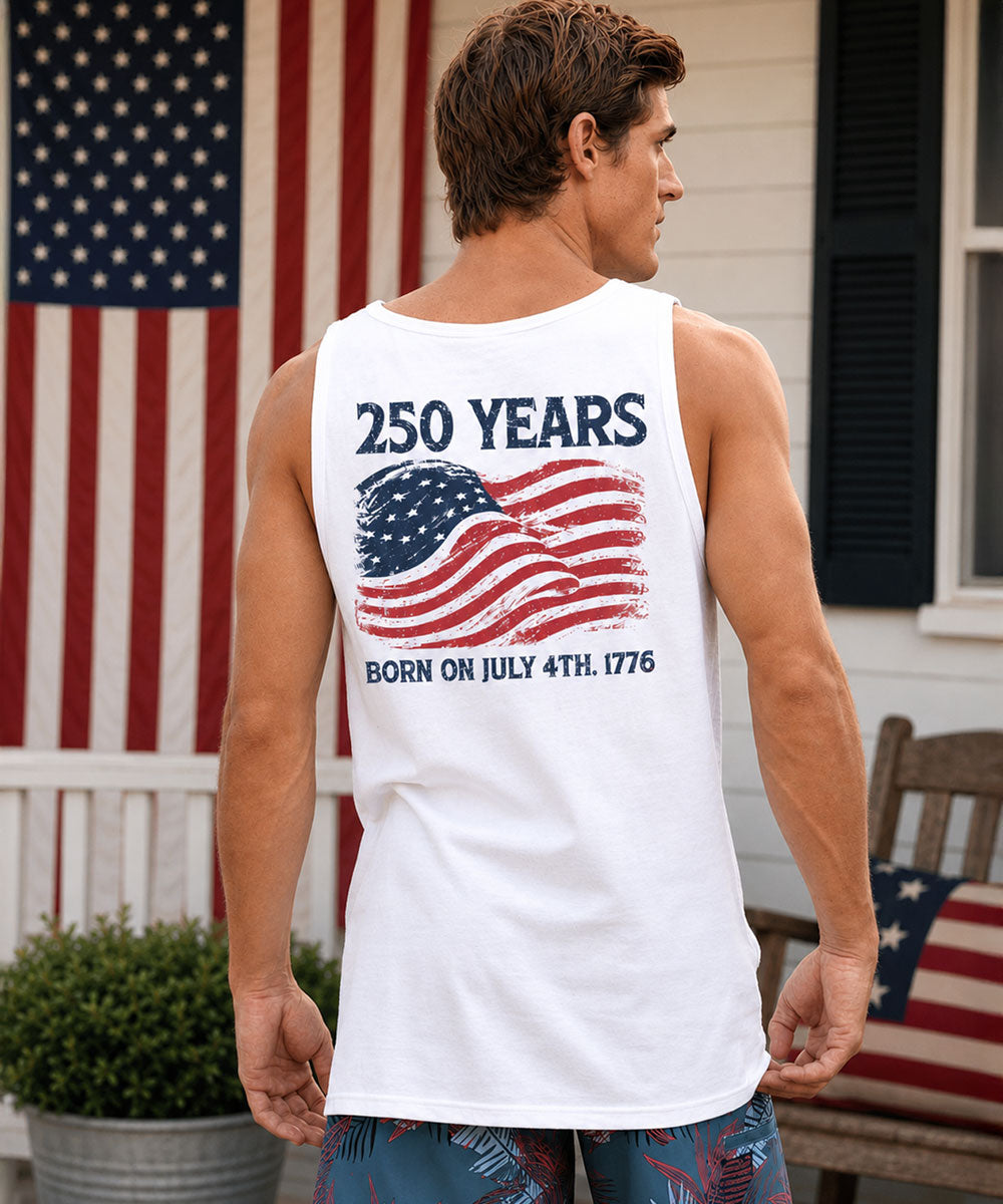 Man wearing a white tank top with American flag design and text, standing in front of an American flag. View 2