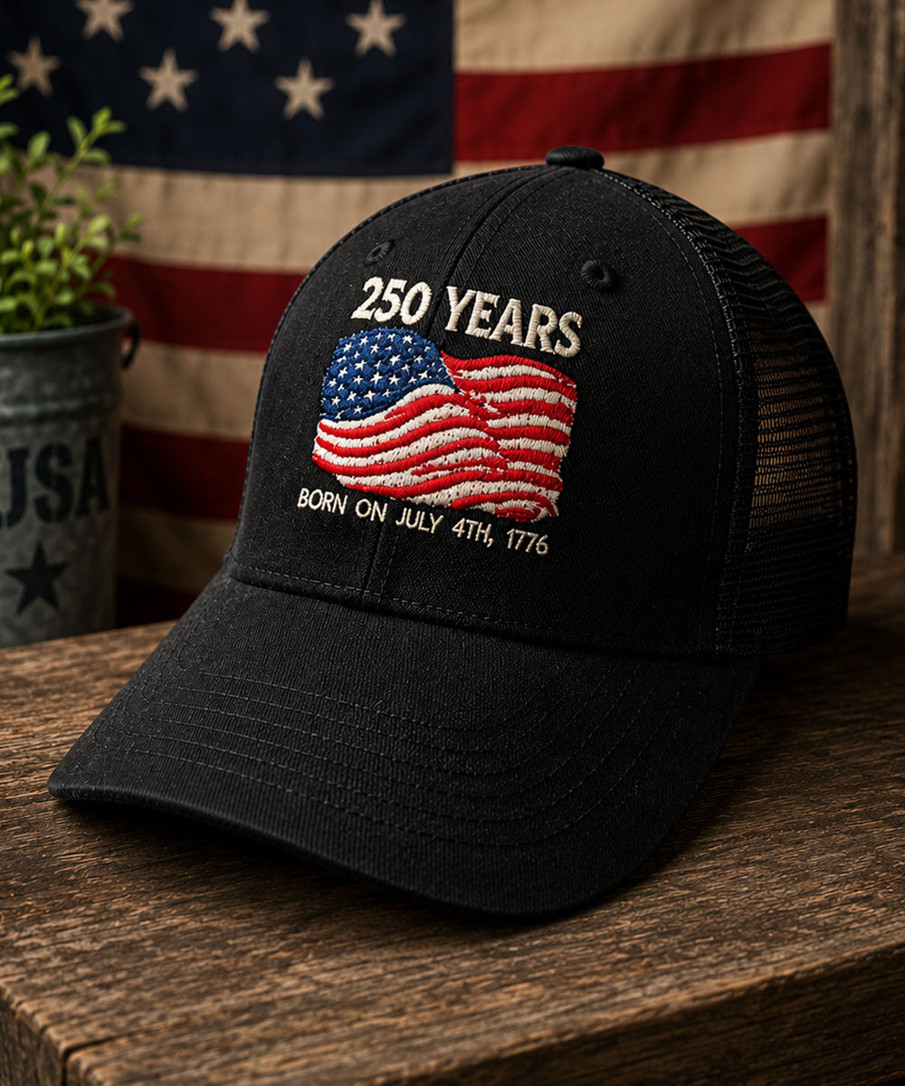 Black cap with '250 Years' and American flag design on a wooden surface with an American flag in the background.