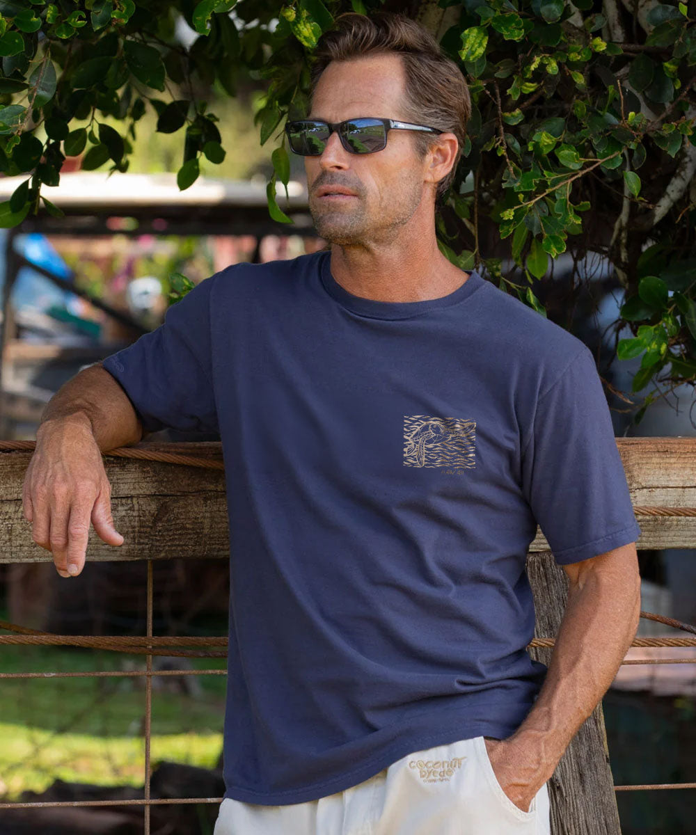 A man wearing a navy blue crewneck t-shirt with a turtle graphic on the left chest, leaning against a wooden fence outdoors under green foliage. View 3