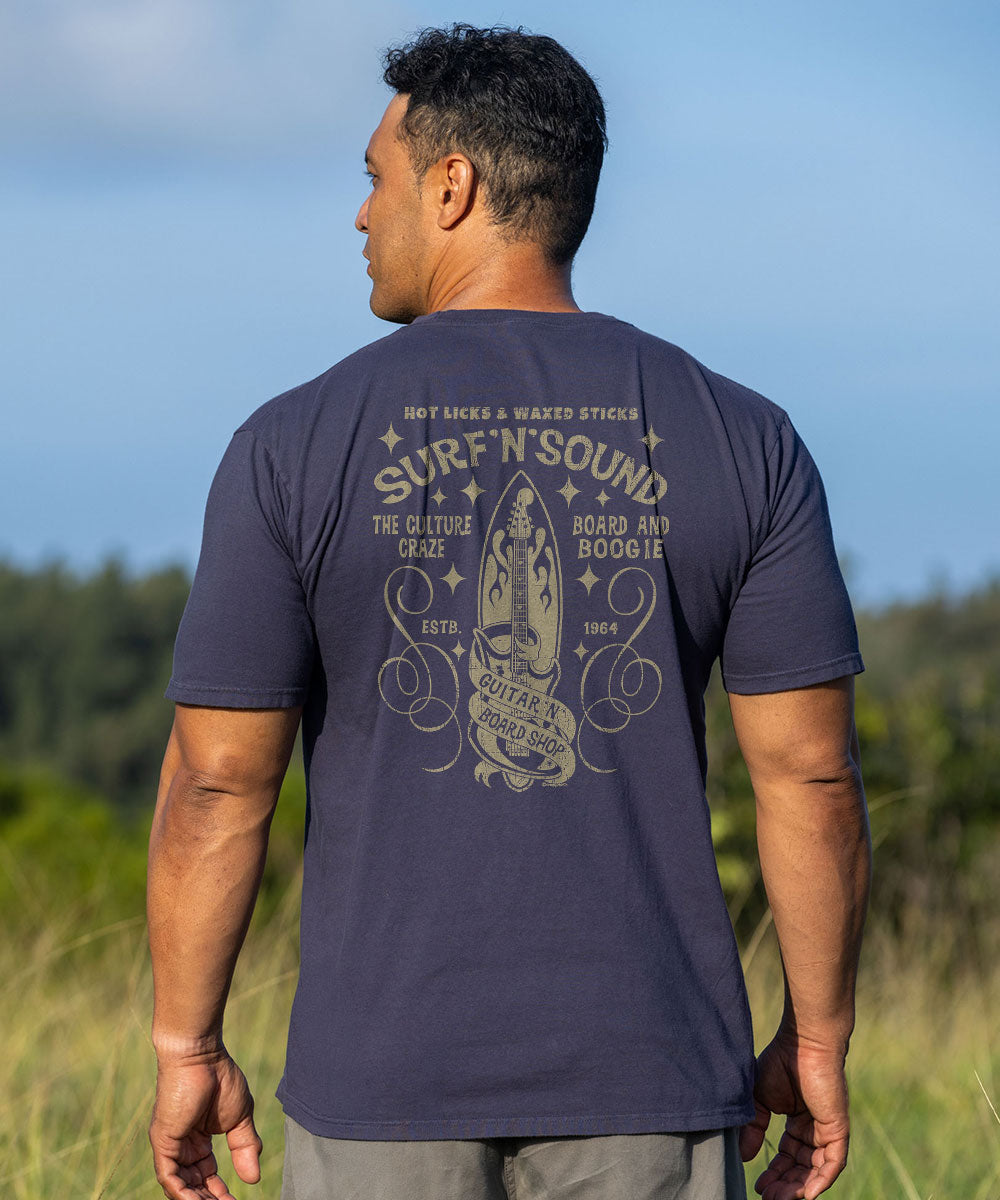 A man wearing a navy blue t-shirt with a large beige graphic print on the back, photographed outdoors against a blurred background of sky and grass. View 2
