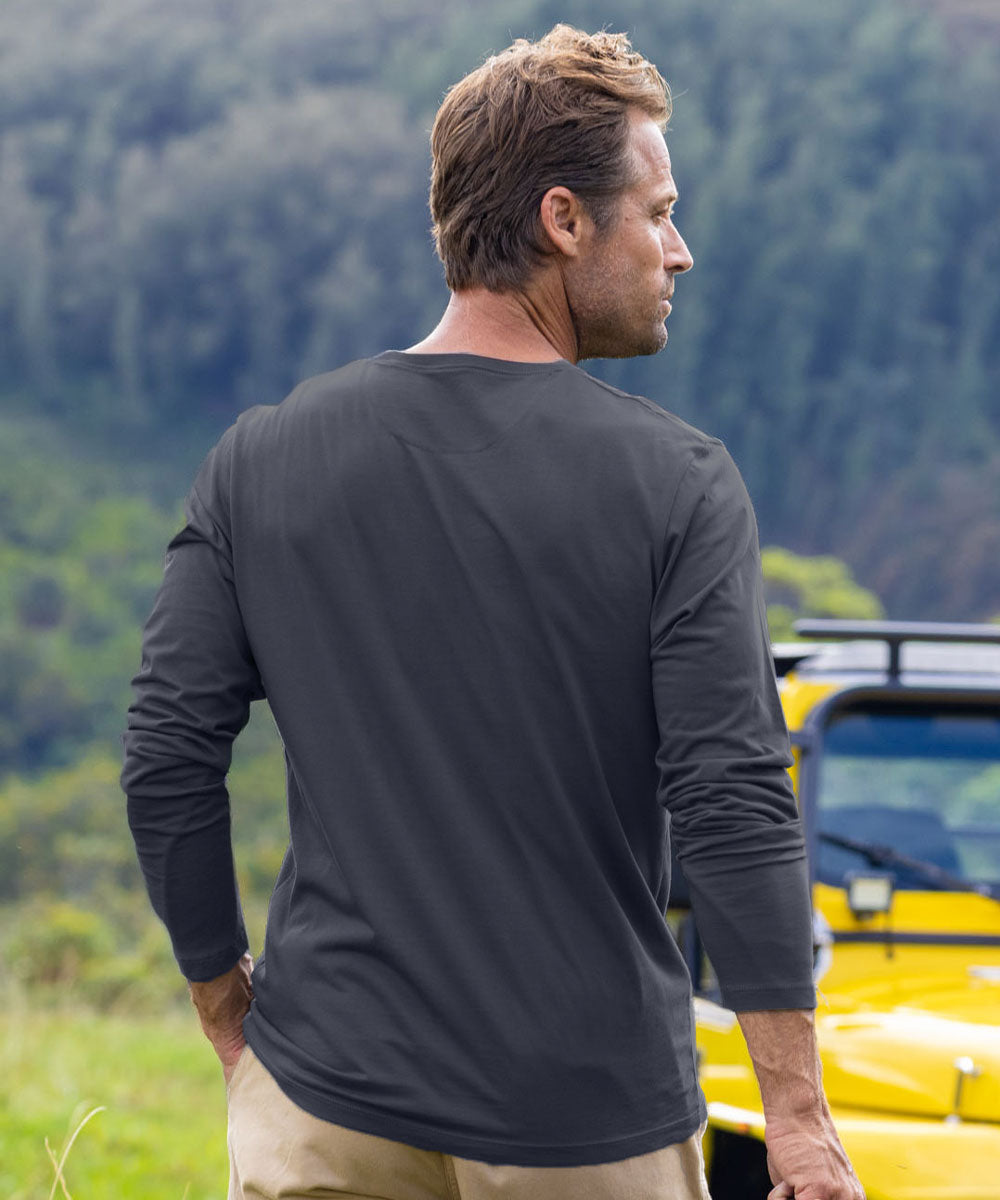 A man wearing a dark charcoal gray long-sleeve performance shirt, shown from the back and side profile. He is standing outdoors in a grassy field with a forested hill in the background and a yellow vehicle partially visible on the right. View 3