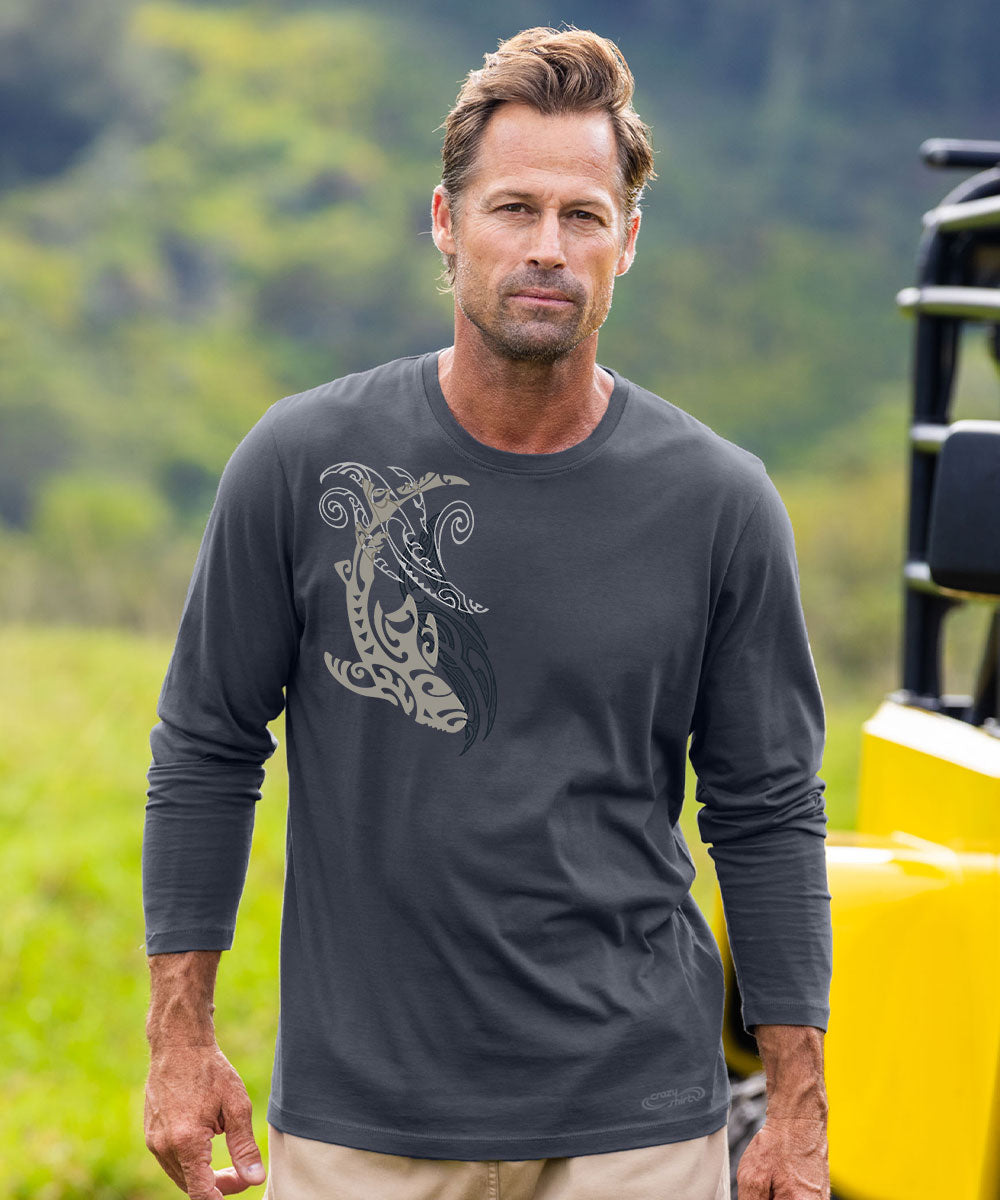 A man wearing a dark gray long-sleeve t-shirt with a light gray Polynesian tribal graphic on the front, standing outdoors in front of a blurred green landscape and a yellow vehicle. View 2