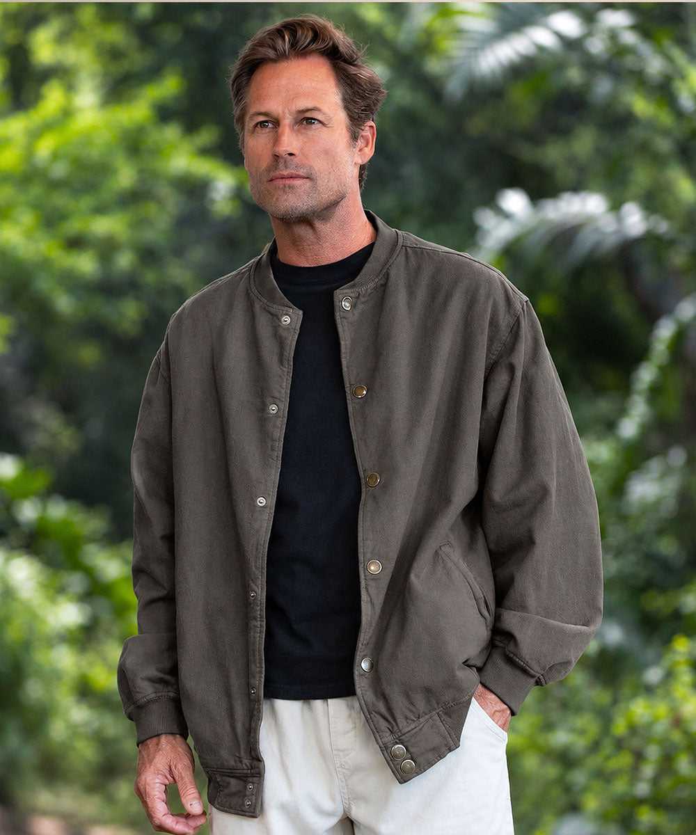 A man wearing a muted olive green bomber jacket with a distressed American flag patch on the chest pocket, layered over a black t-shirt and cream pants, standing against a background of foliage View 3
