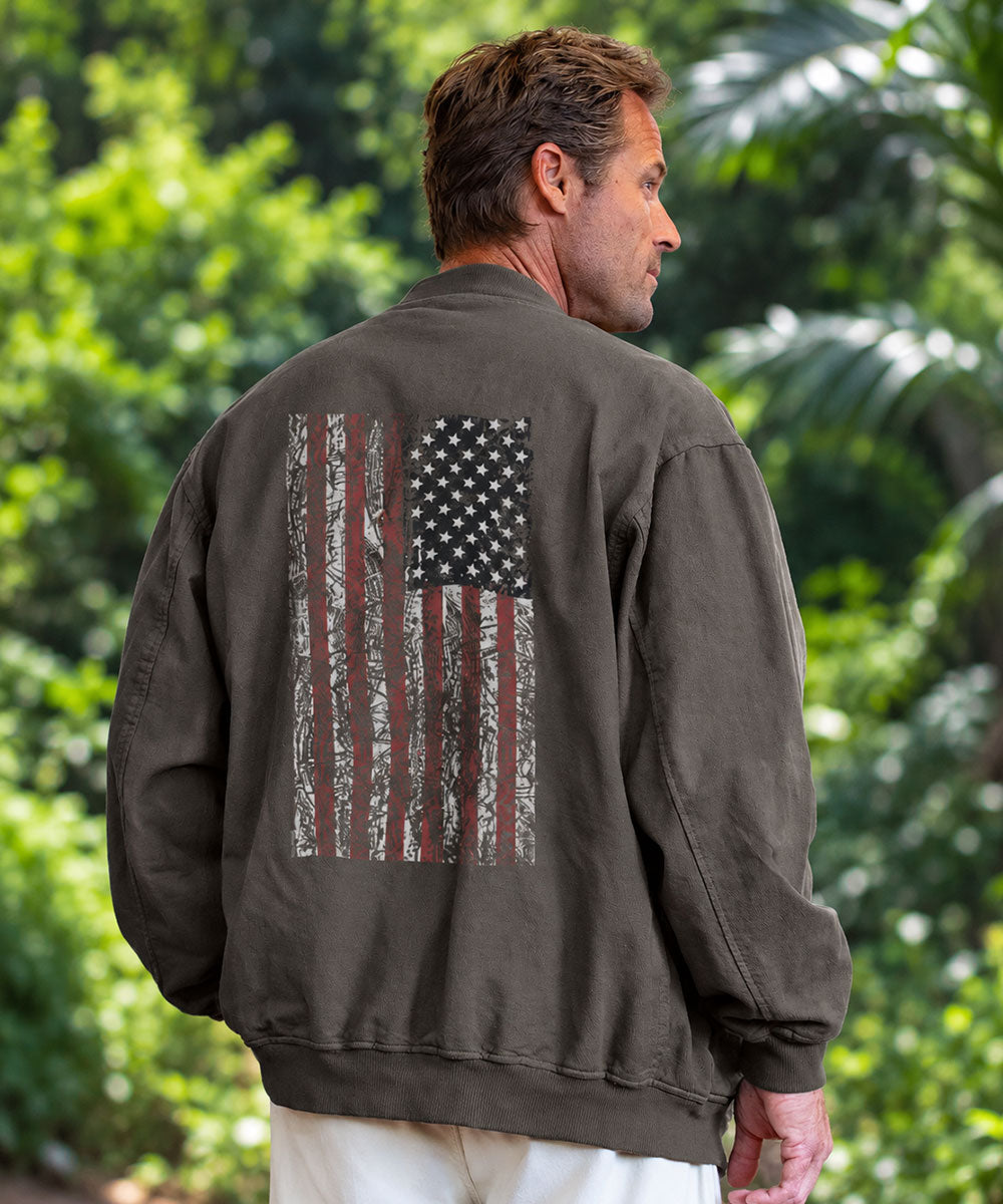 A man wearing a dark olive brown bomber jacket, viewed from behind to show a distressed American flag graphic printed on the back. The jacket is olive brown with a faded, distressed vertical American flag design on the back in black, gray, and muted red. View 2