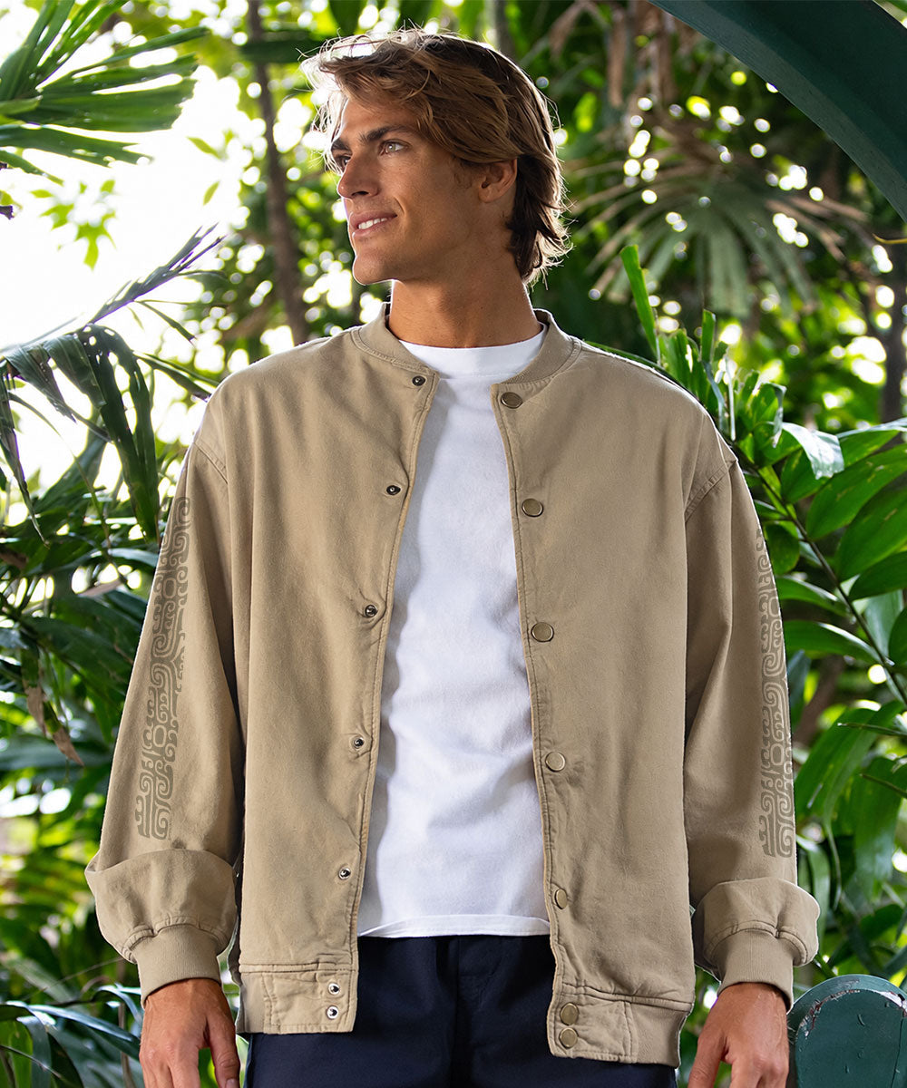A man wearing a beige button-front jacket with decorative sleeve embroidery over a white t-shirt, standing outdoors in front of lush green foliage. View 2