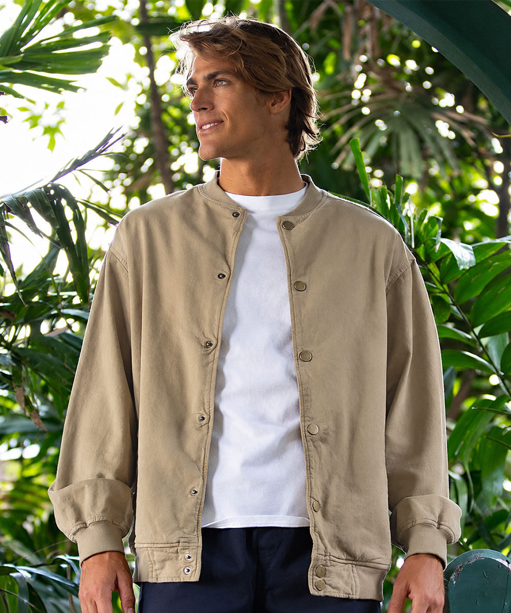 A man wearing a beige button-front jacket over a white t-shirt and dark pants, standing outdoors in front of lush green tropical foliage. View 2