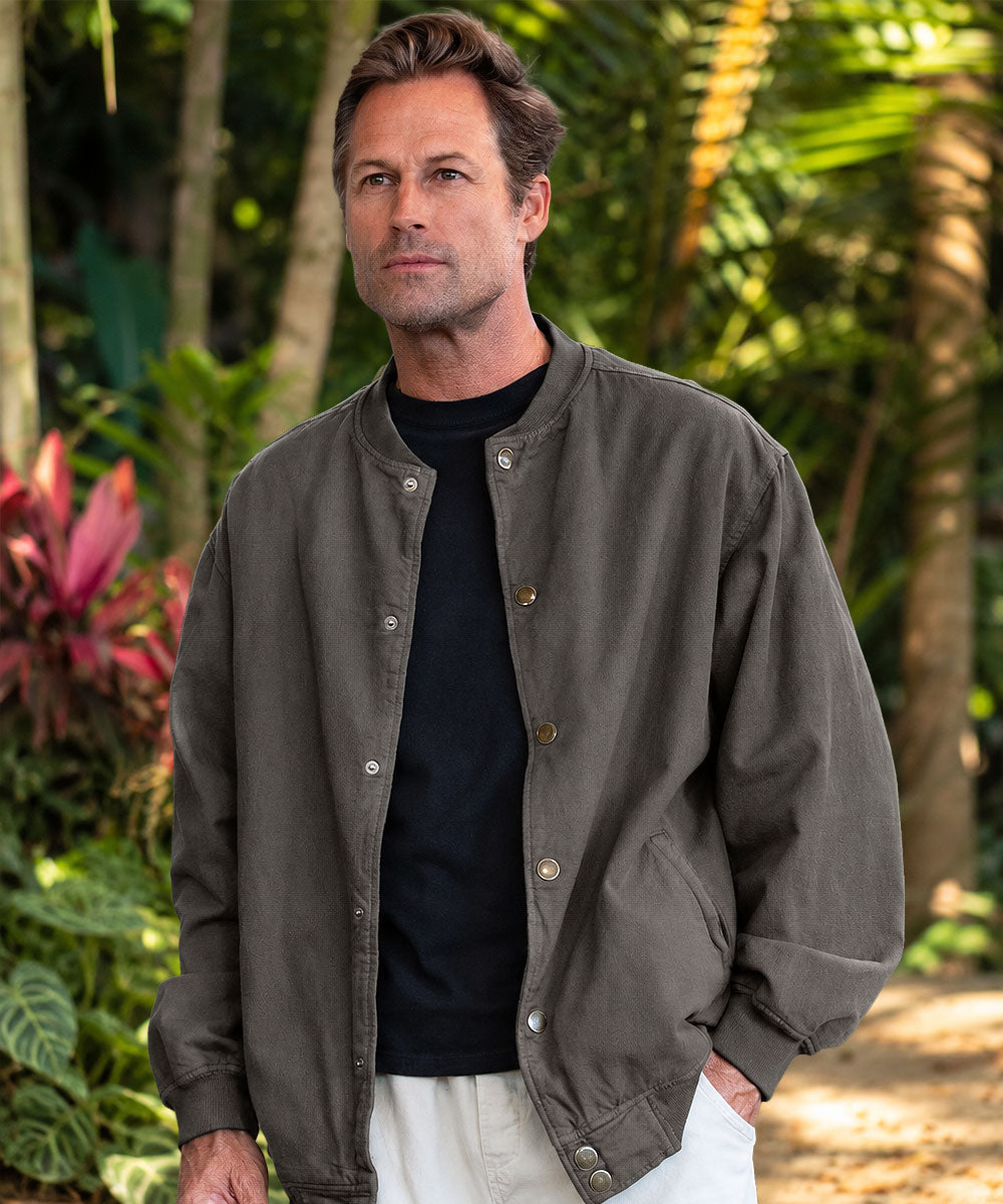 A man wearing a dark gray bomber jacket over a black t-shirt and off-white pants, standing in front of foliage