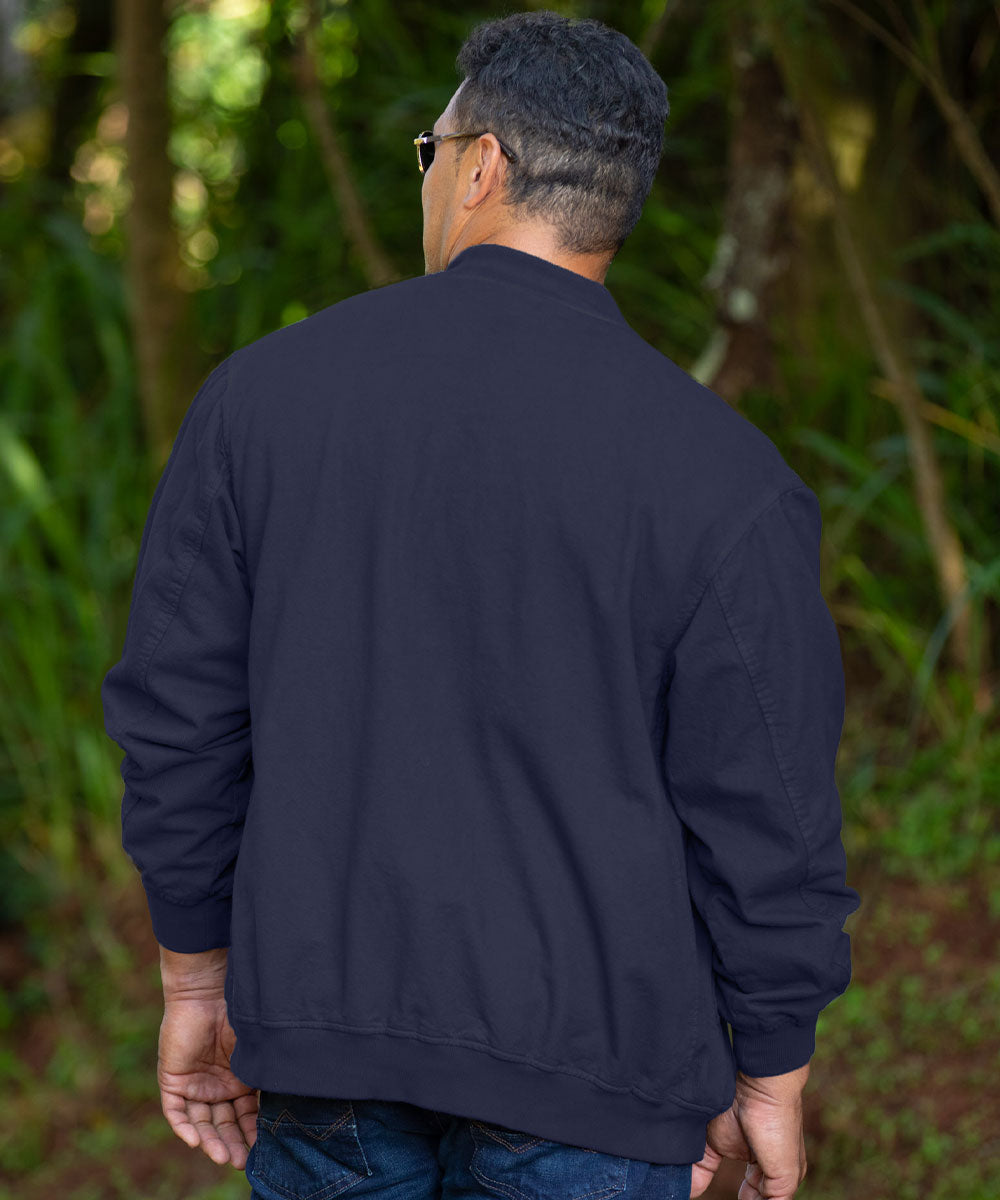 A man viewed from behind wearing a dark navy blue bomber jacket and blue jeans, standing outdoors in a lush green forest setting. View 3