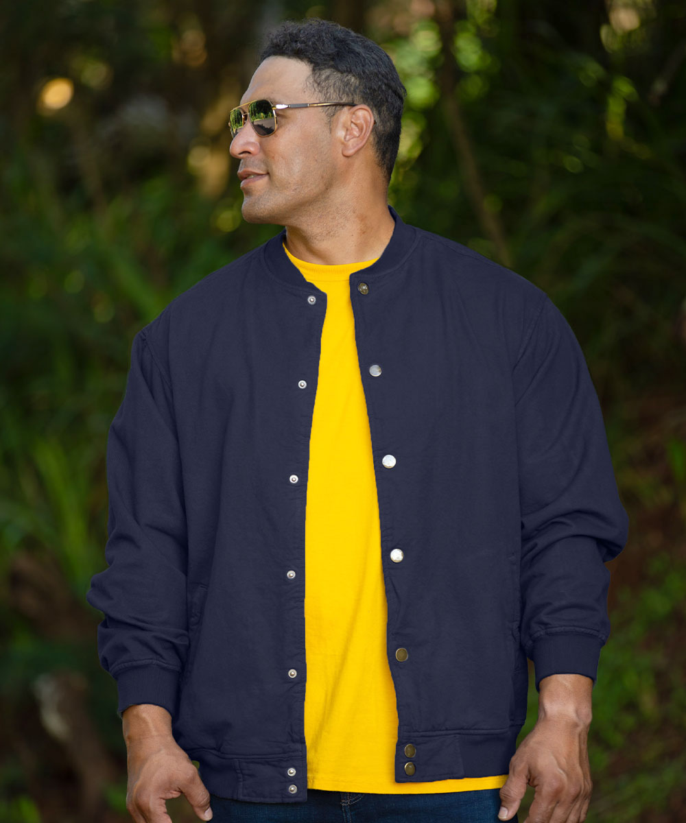 A man wearing a navy blue button-up varsity-style jacket over a bright yellow t-shirt, standing outdoors against a blurred green forest background. View 2