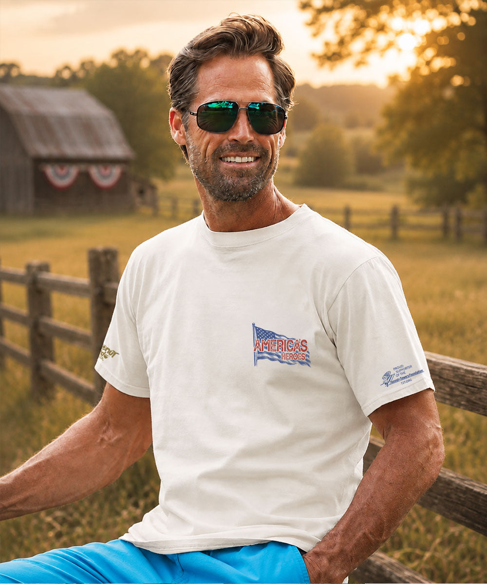A man wearing a cream-colored t-shirt with a patriotic 'america's heroes' graphic on the left chest, paired with blue pants and green-tinted sunglasses, set against a farm background. View 3
