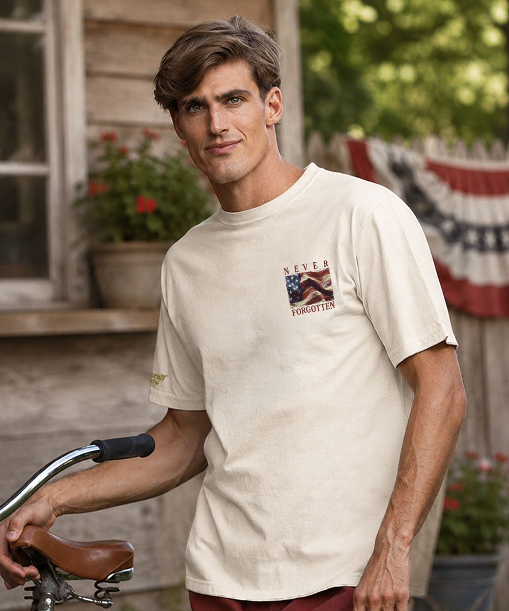 A man wearing a cream-colored short-sleeve t-shirt with a 'Never Forgotten' graphic on the left chest, standing outdoors in front of a wooden house and holding a bicycle handlebar. View 3