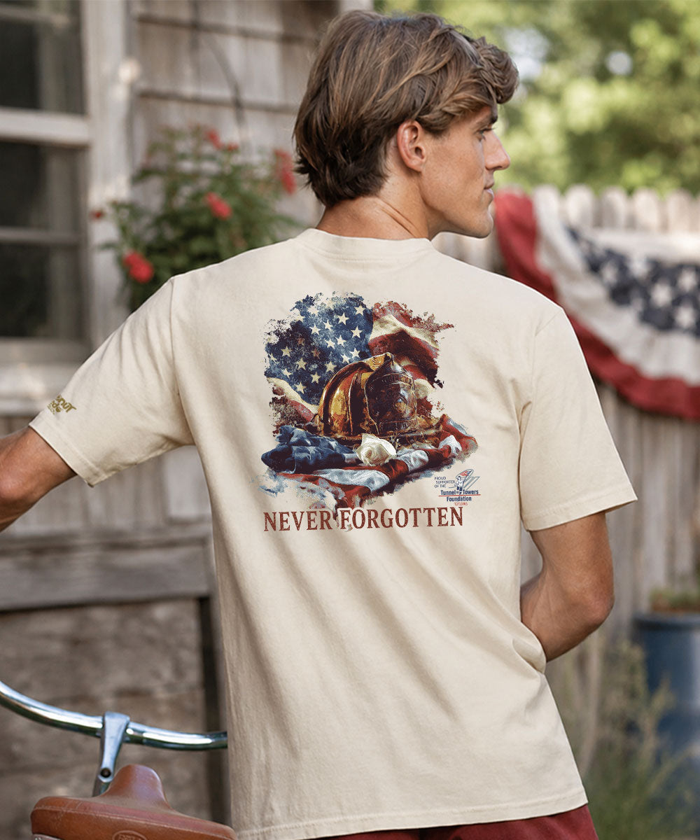 A man wearing a beige t-shirt with a large 'Never Forgotten' graphic on the back, standing outdoors in front of a wooden house View 2