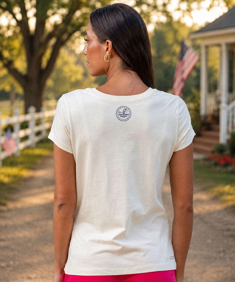 A woman viewed from behind wearing a cream-colored short-sleeve t-shirt with a circular logo on the upper back, set against a blurred outdoor background View 3