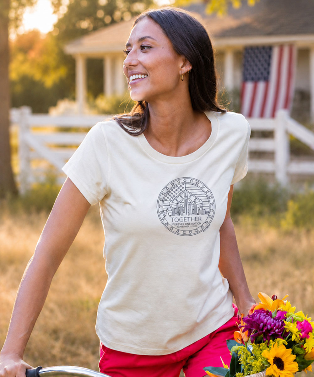 A woman riding a bicycle outdoors wearing a cream-colored t-shirt with a gray circular graphic on the chest that reads 'TOGETHER' with 'United States Army' and '101st Training Command' written below. She is also wearing bright pink pants and holding a bouquet of colorful flowers. View 2