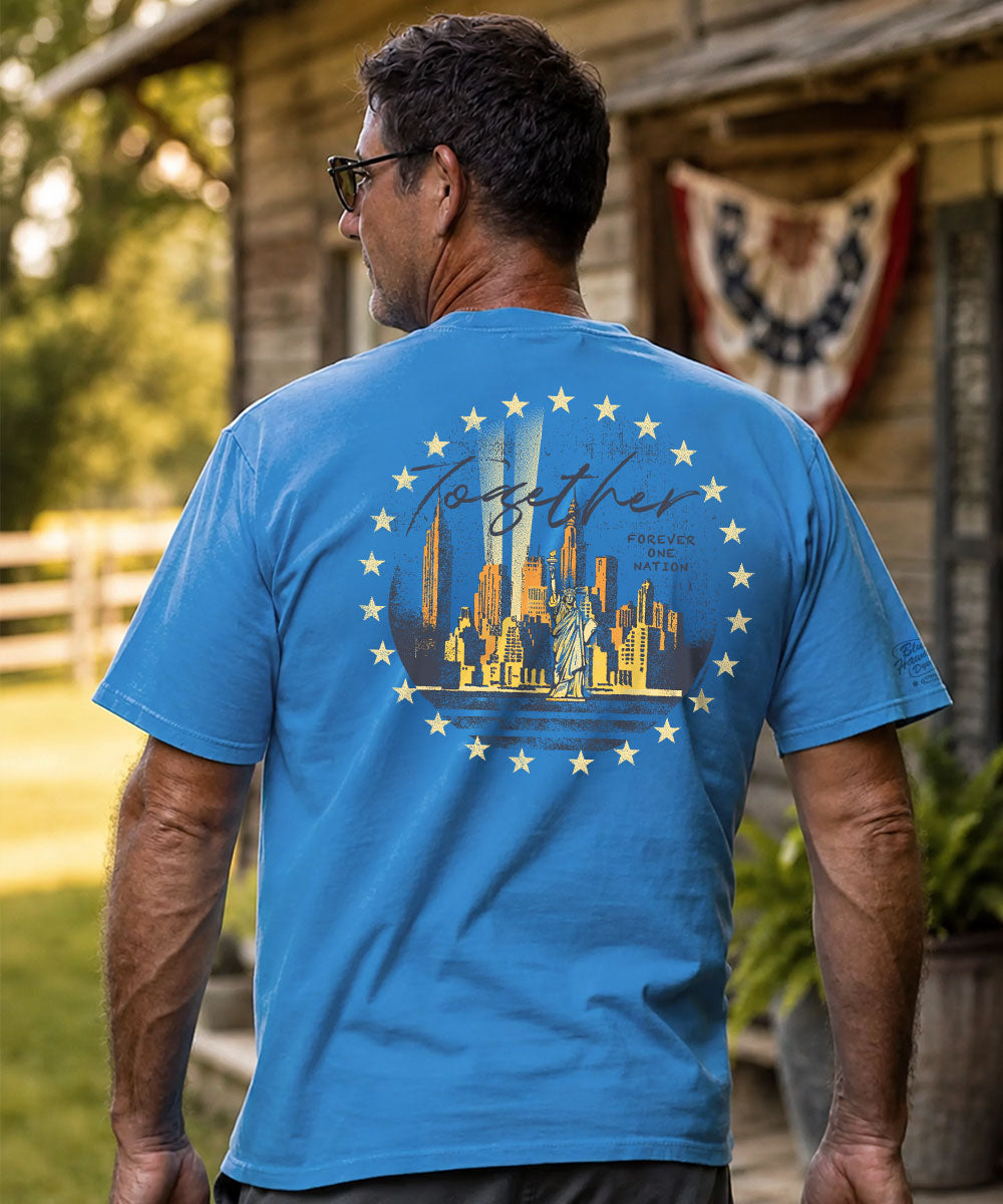 A man viewed from behind wearing a bright blue t-shirt with a large graphic on the back. The graphic features a golden city skyline, the Statue of Liberty, and a circle of stars surrounding the word 'Together' in script. View 2