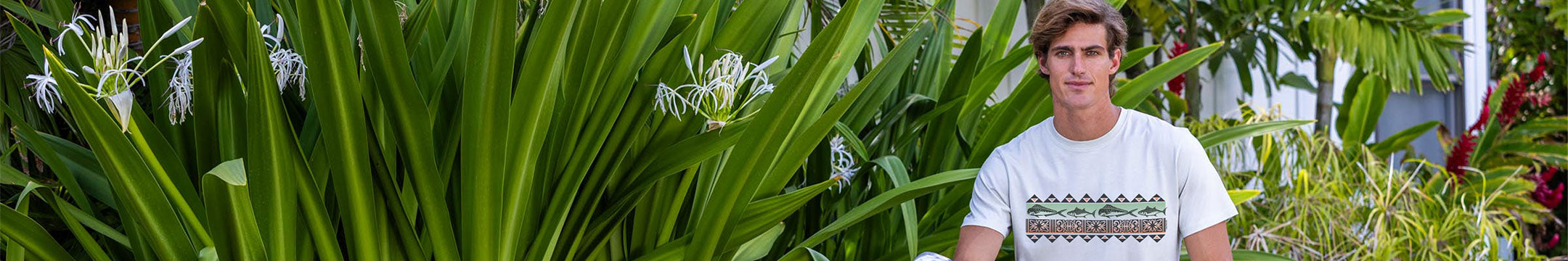 Man wearing a white t-shirt with a pattern, standing among green plants.