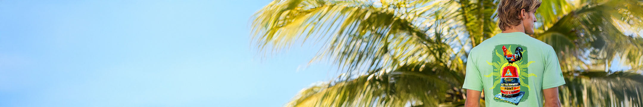 Person wearing a green t-shirt with a colorful design, standing under a palm tree with a clear blue sky.