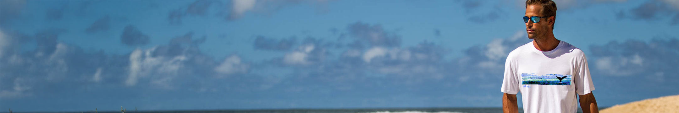 Man wearing a white t-shirt with a logo standing on a beach with blue sky and clouds in the background
