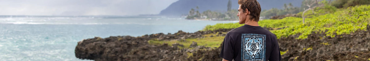 A man stands on a rocky coastline looking out toward the ocean, wearing a black t-shirt with a blue and white graphic print on the back.