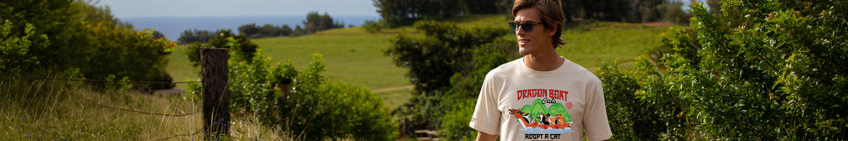 A man wearing a white graphic t-shirt with a colorful cartoon design stands outdoors in a grassy field with trees in the background.