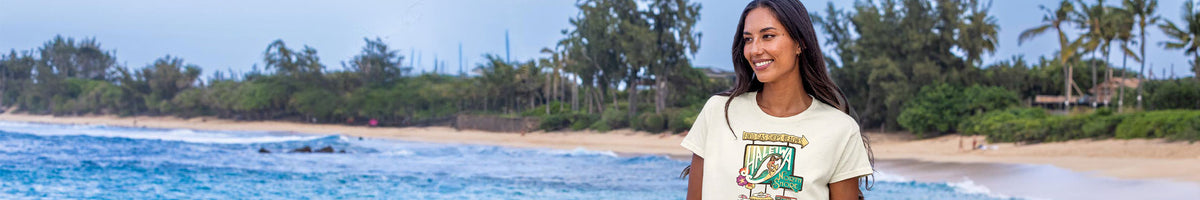 A woman standing on a beach wearing a cream-colored t-shirt with a green and yellow graphic print. The background features a sandy beach, blue ocean water, and a line of green palm trees under a bright sky.