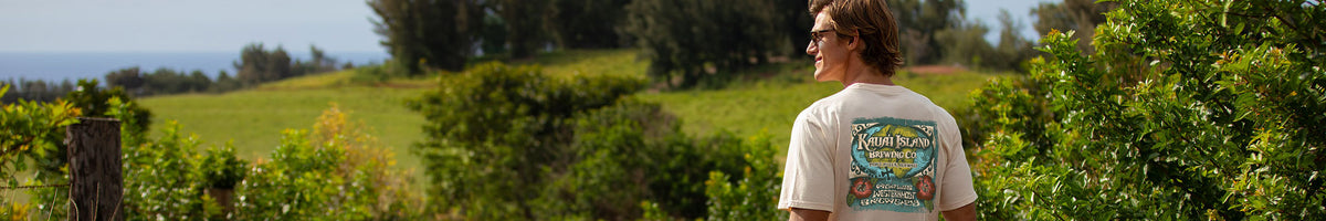 A man wearing a white graphic t-shirt stands outdoors in front of a lush green landscape, looking to the left. The back of the t-shirt features a colorful graphic.