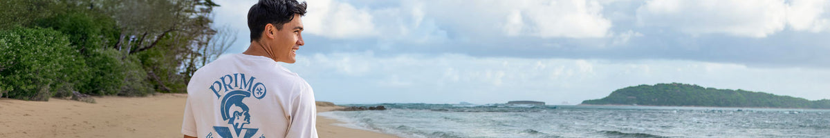 A man wearing a white short-sleeve t-shirt with a blue graphic print on the back stands on a sandy beach looking out toward the ocean under a cloudy sky.