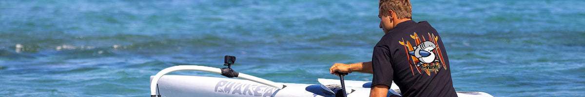 A man wearing a black t-shirt with a large orange and white graphic on the back sits on a white boat in the blue ocean water.