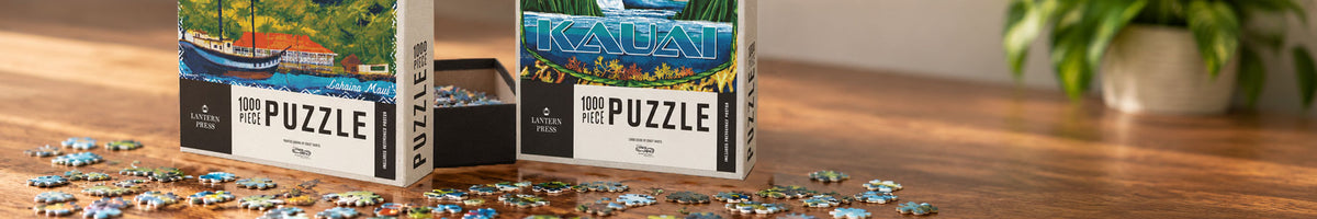Two jigsaw puzzle boxes labeled Kauai and Maui sit on a wooden table with loose puzzle pieces scattered around. A blurred potted plant appears in the background.
