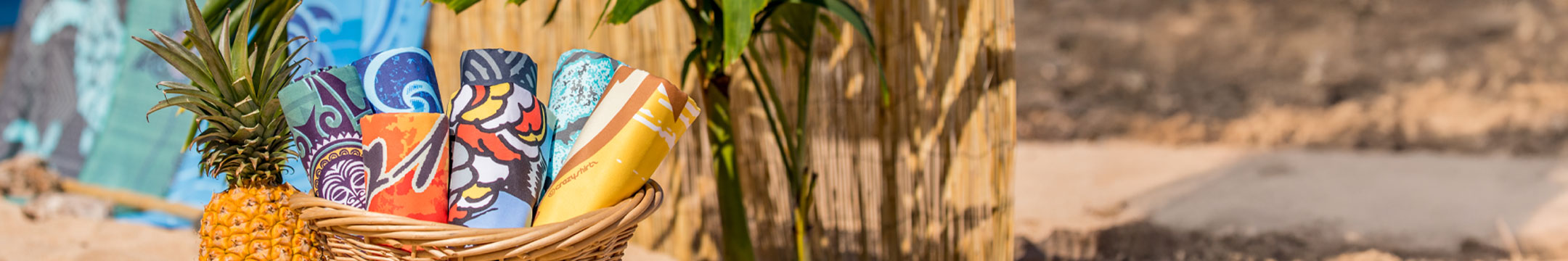 Basket with tropical-themed towels including a pineapple, and a colorful box on a sandy background.