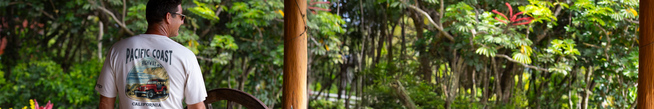 Person wearing a 'Pacifc Coast' t-shirt in a tropical setting with lush greenery.
