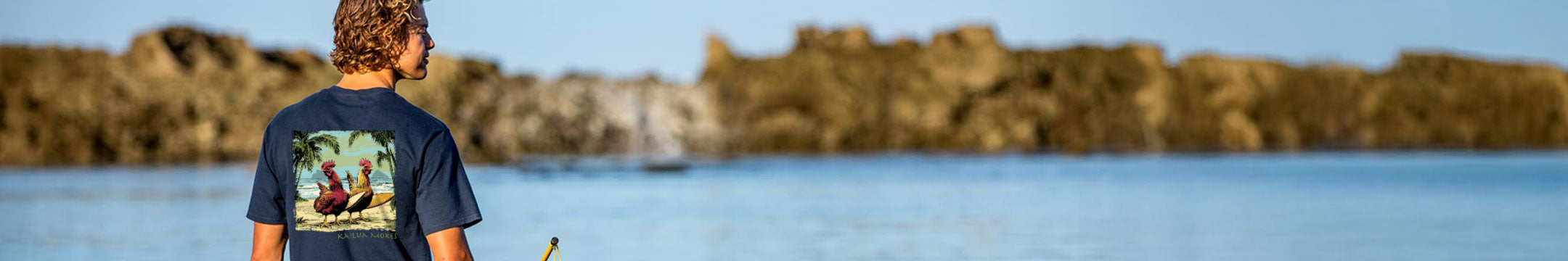 Person wearing a Crazy Shirt Navy Tee, standing by a body of water with rocky cliffs in the background.