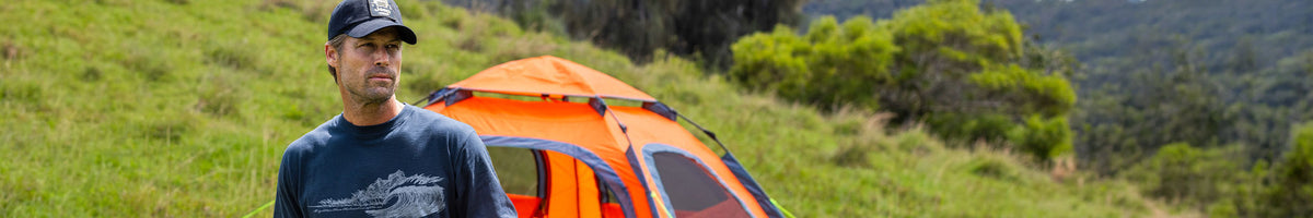 A man wearing a black baseball cap and a dark graphic t-shirt stands outdoors in a grassy mountain setting next to an orange and blue camping tent.