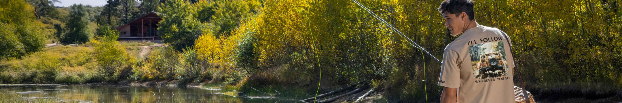 Man fishing by a river with trees and a covered bridge in the background
