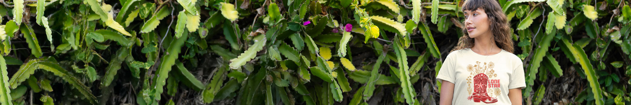 woman standing in front of a cactus wall wearing a tan shirt with a boot on it