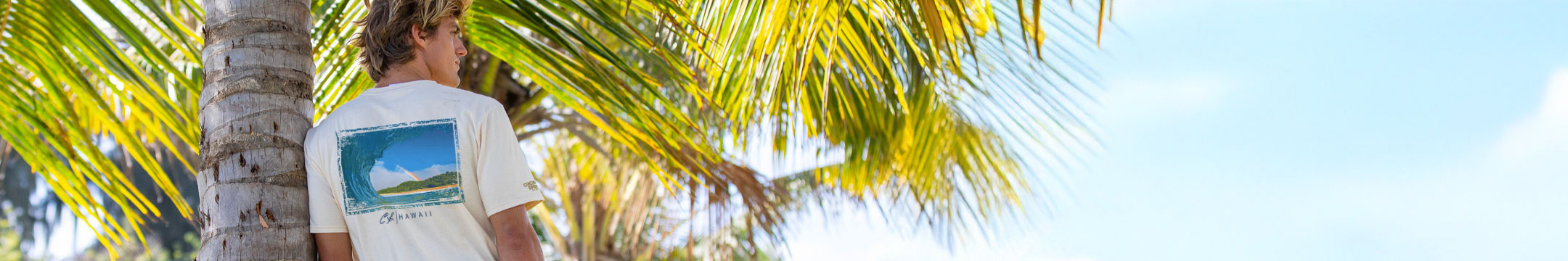 Person wearing a white t-shirt with a graphic design, standing under palm trees with a clear blue sky.
