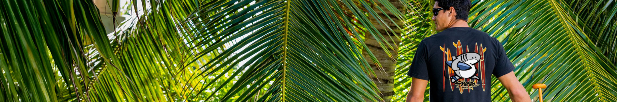 Person walking through a tropical setting with palm leaves wearing a Sharka Tee