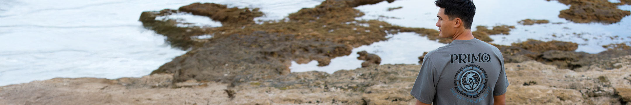 Person wearing a 'PRIMO' shirt standing on a rocky shoreline.